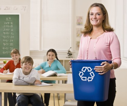 A smiling woman with shoulder-length light brown hair, wearing a pink cardigan, stands in a classroom holding a blue recycling bin with a white recycling symbol printed on it. Behind her, three children are seated at light wooden desks, attentively looking towards her, with books and notebooks open in front of them. The classroom features light-colored walls, a green chalkboard with handwritten math calculations on the left, and windows with white blinds that allow natural light to fill the room. The scene depicts an educational setting focused on recycling awareness, subtly connecting to rubbish management services provided by Commercial Waste Purley, located in the Croydon area near Purley postcode region. The environment appears tidy and well-organized, with the children engaging in the lesson while the woman prepares for a waste reduction activity, emphasizing the importance of responsible rubbish disposal and recycling education.