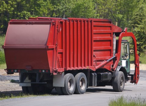 A large red industrial waste collection truck parked on a paved driveway with a background of green trees and foliage. The truck features a rectangular, open-top metal container with vertical reinforcement ribs and a smooth, painted surface. The container is designed for rubbish removal and is positioned behind the cab, which is white with black tires and grey wheel rims. The environment appears to be outdoors with natural daylight illuminating the scene, indicating a rural or semi-urban setting near Purley, as suggested by the context of commercial waste services in the area. This vehicle is associated with rubbish collection and waste management services, similar to those provided by Commercial Waste Purley, and is suited for bulk waste disposal, possibly for a commercial or industrial client in the local area.