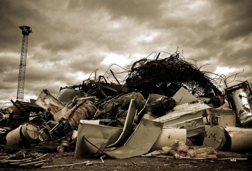A large pile of scrap metal and debris situated outdoors on a paved surface, with various discarded items including twisted metal beams, broken appliances, and damaged vehicle parts. The materials appear weathered, with some surfaces rusted and oxidised, exhibiting shades of grey, beige, and metallic tones. At the top of the heap, there is a tangled mass of dark, dense wire or cable. The background features a cloudy sky with an overcast atmosphere, and a tall metallic structure, possibly a crane or tower, positioned on the left side of the image. The scene is professional and neutral, typical of waste collection or rubbish removal operations in a commercial setting, consistent with the services provided by Commercial Waste Purley near Croydon, with the debris arrangement indicating an organized disposal process on a paved area suitable for such activity.