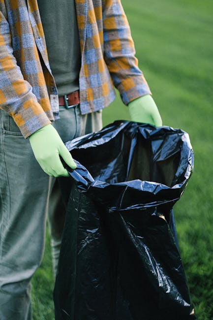 A person wearing a checked shirt in shades of yellow, brown, and gray, along with green rubber gloves, is holding a large black rubbish bag that is partially open, revealing its empty interior. The in