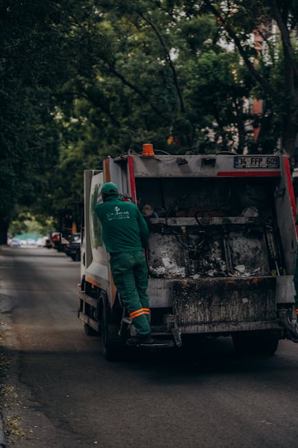 A person wearing a checked shirt in shades of yellow, brown, and gray, along with green rubber gloves, is holding a large black rubbish bag that is partially open, revealing its empty interior. The in